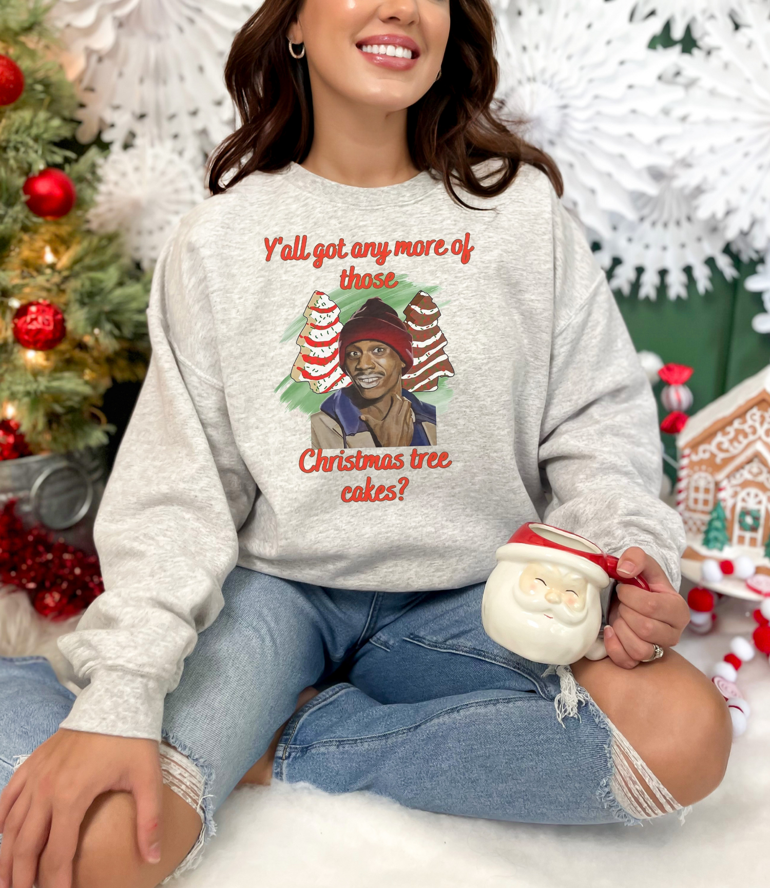 A person wearing a gray sweatshirt with an image of Tyrone Biggums from Dave Chappelle Show, alongside a Christmas tree cake, holding a mug, with a festive background.